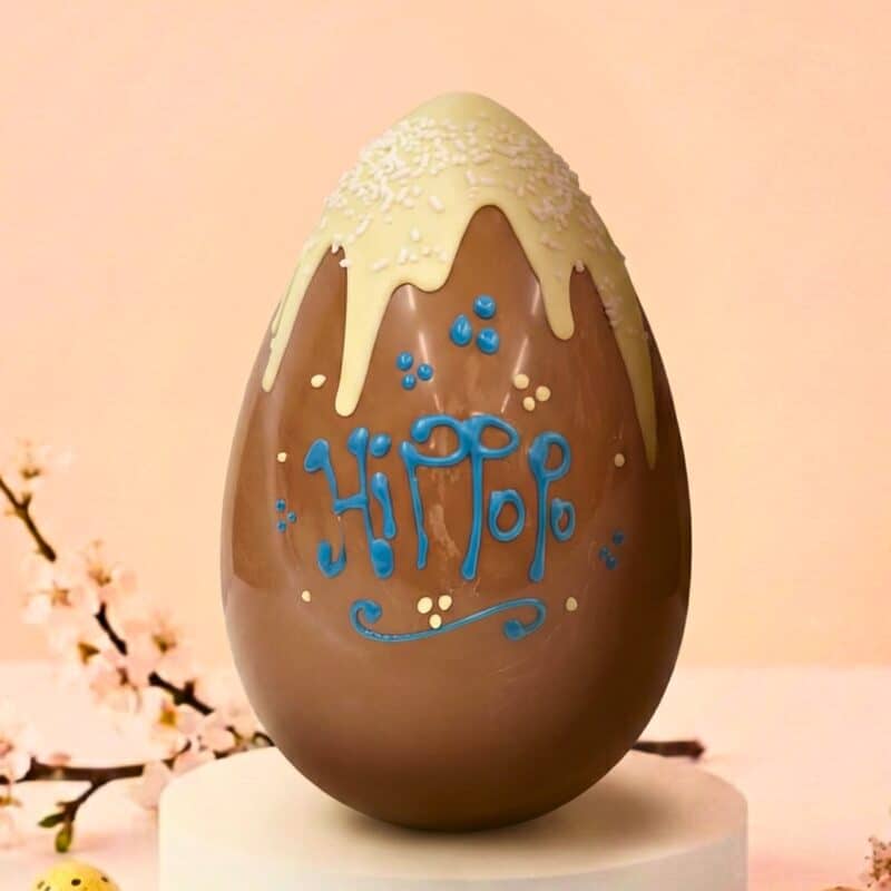 Uovo di Pasqua artigianale in cioccolato al latte, decorato con colata bianca, codette di zucchero e scritta Hippopo a mano in azzurro. Sfondo primaverile.