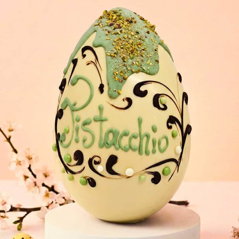 Uovo di Pasqua artigianale in cioccolato bianco, decorato con colata verde al pistacchio, granella croccante, eleganti ghirigori in cioccolato fondente e scritta Pistacchio a mano. Sfondo primaverile.