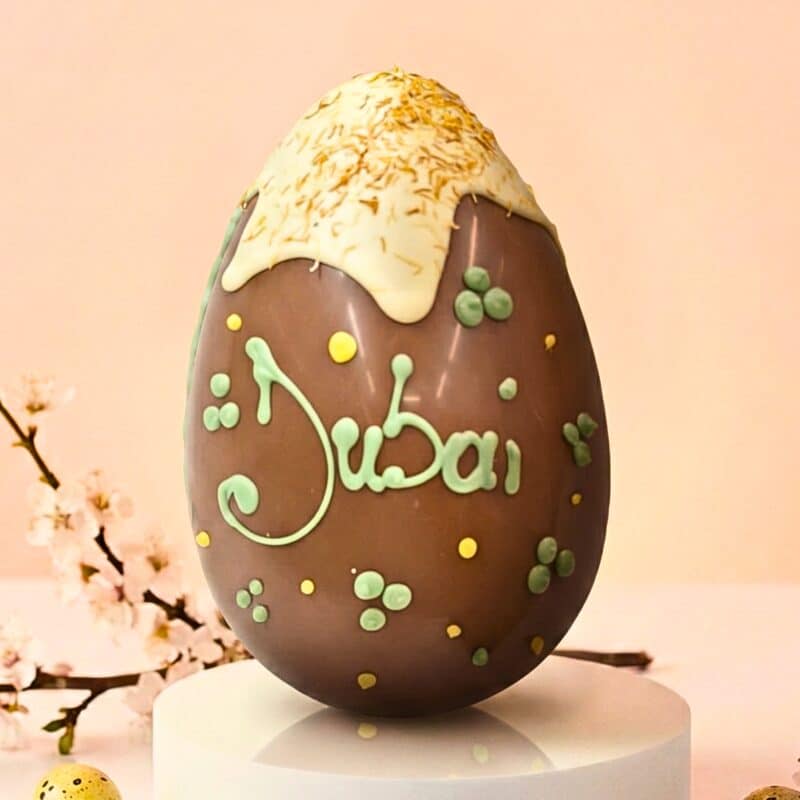 Uovo di Pasqua artigianale in cioccolato al latte, ispirato al trend Dubai chocolate. Decorato con colata chiara, pasta kataifi croccante, pois e scritta Dubai a mano in verde pistacchio. Sfondo primaverile.