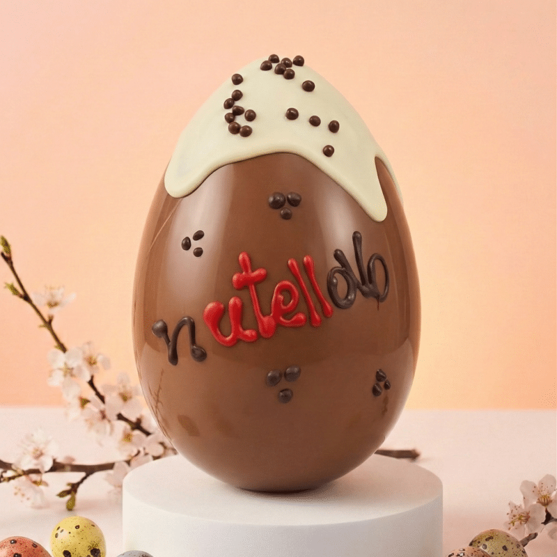 Grande uovo di Pasqua artigianale Nutellotto da 700g, cioccolato al latte con colata di gianduia e granella di nocciole, decorazioni fatte a mano, su piedistallo bianco.